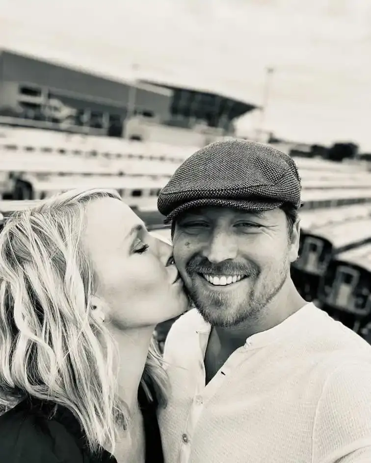 Black and white selfie of Kelli Giddish kissing her smiling husband, Beau Richards, on the cheek, with a stadium or train yard in the background.