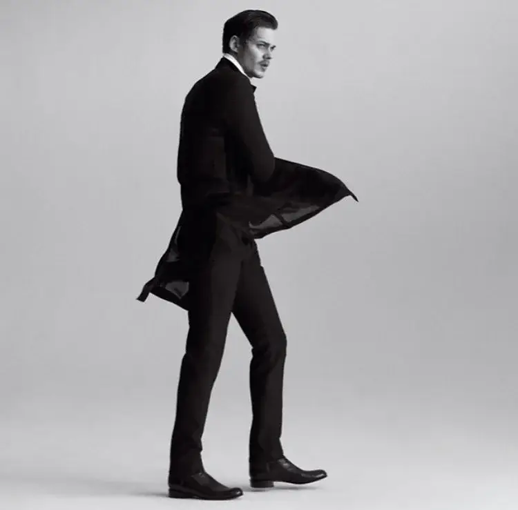 Black and white full-body shot of actor Bill Skarsgård in a slim black suit with a long coat, striding to the side with the coat blowing dramatically.