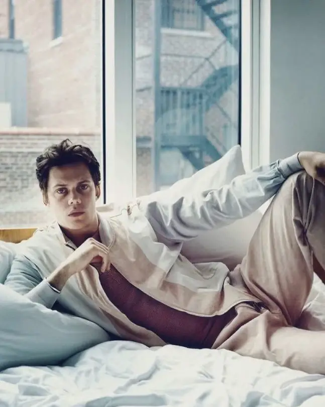 Actor Bill Skarsgård reclines on a white bed in a casual jacket and tan trousers, looking directly at the camera in front of a window.