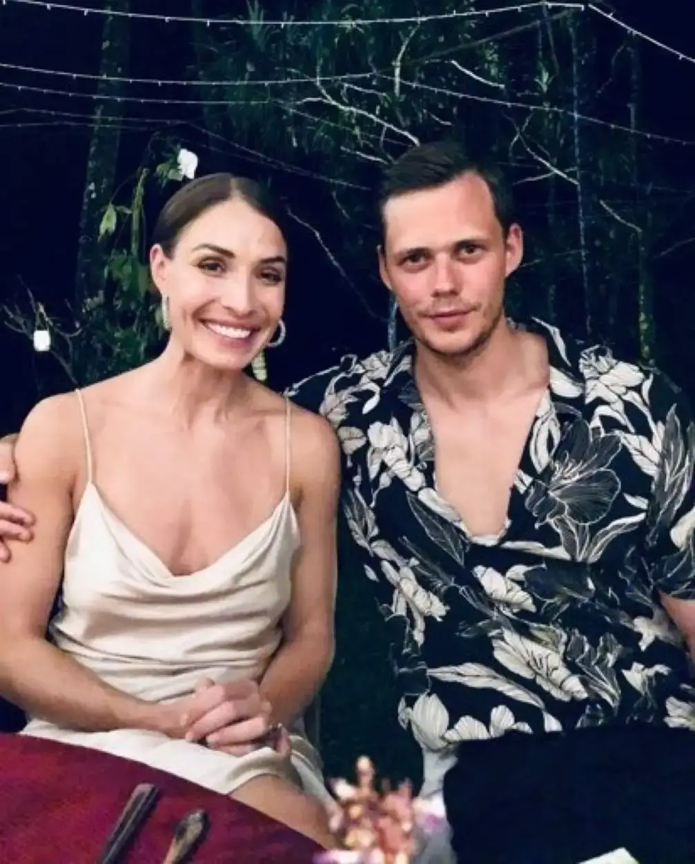 Actor Bill Skarsgård in a floral button-down shirt sits beside a smiling Alida Morberg in a cream silk dress in an event.