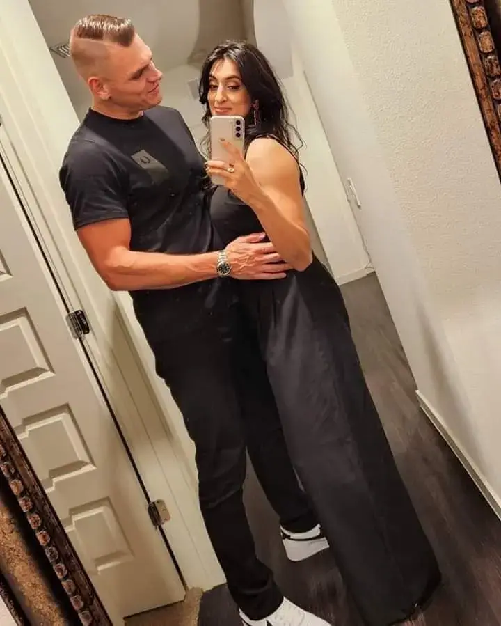 Professional wrestler Gunther stands with his arm around his wife, Jinny Sandhú, as she takes a mirror selfie of them both wearing matching black outfits.
