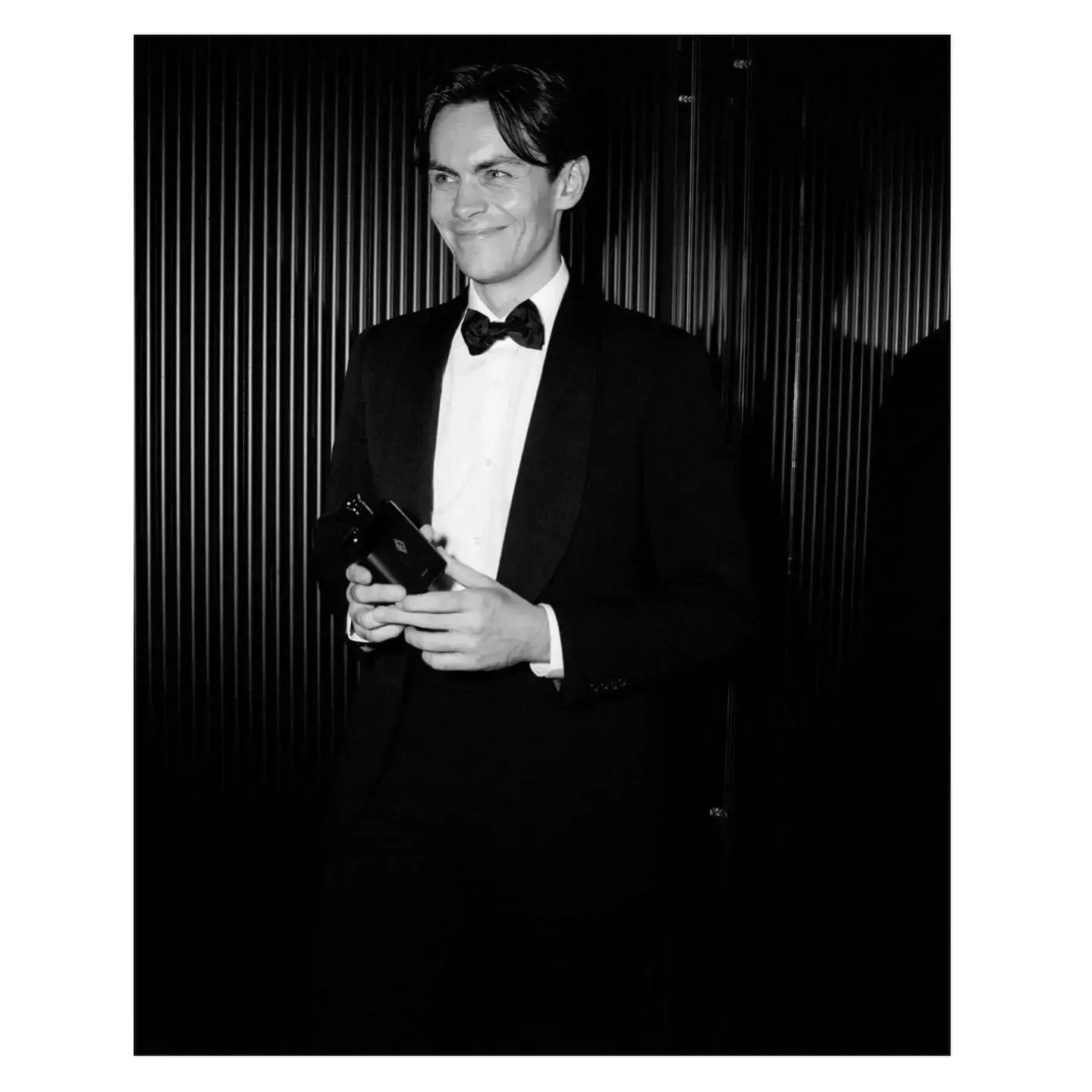 Black and white portrait of actor Freddy Carter smiling in a classic tuxedo and bow tie while holding a small black bottle.