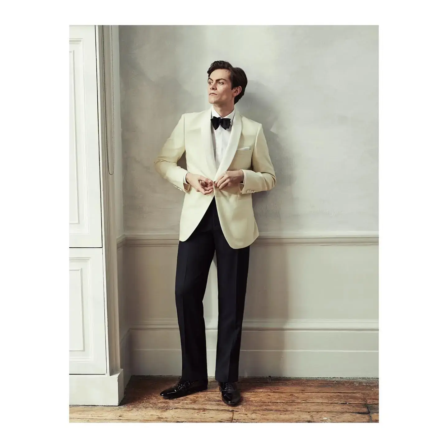 Actor Freddy Carter stands against a neutral wall in a cream-colored tuxedo jacket and black bow tie, looking off to the side while adjusting his jacket buttons.
