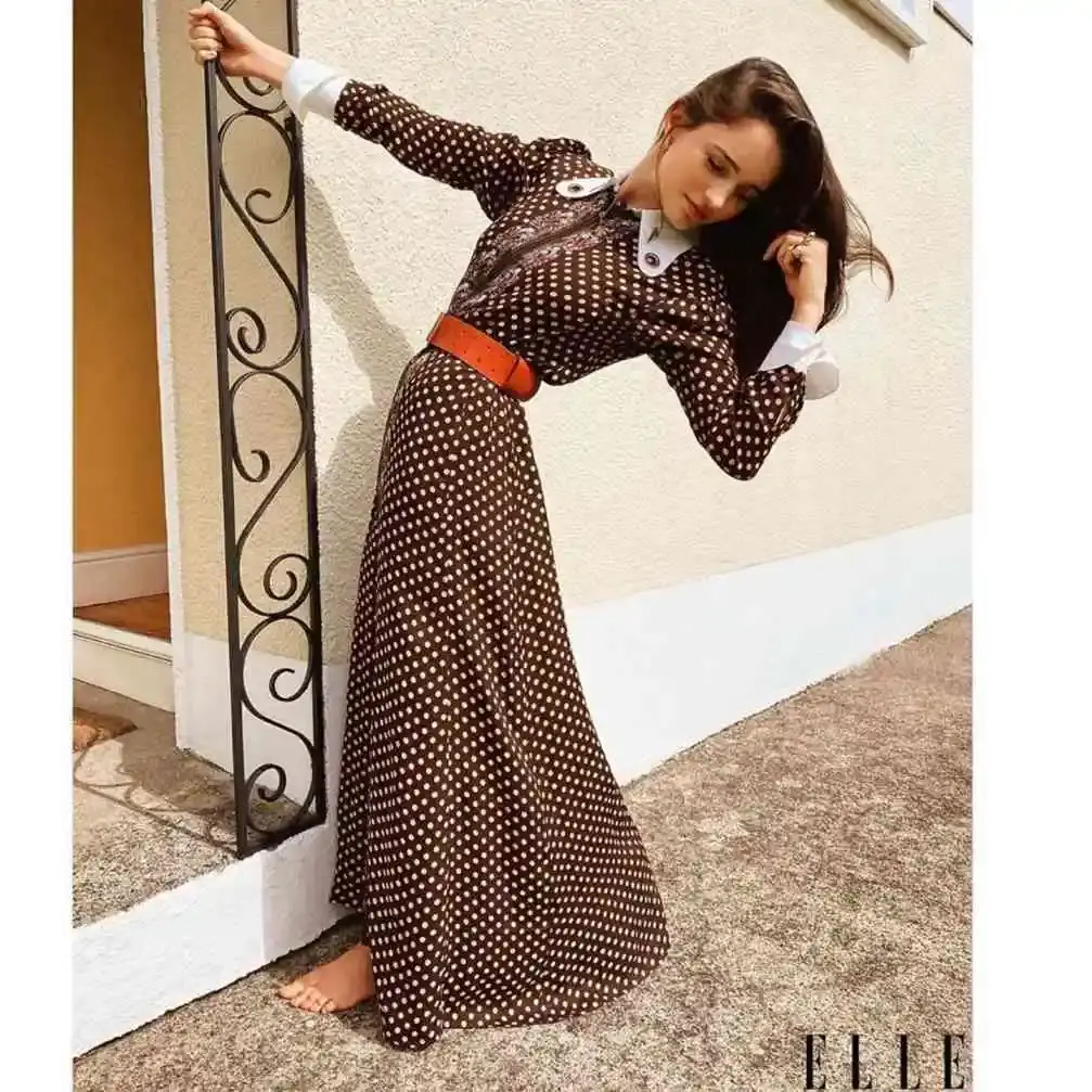 Actress Aisling Franciosi poses barefoot in a long brown polka-dot dress with a bright orange belt while leaning against a iron railing.