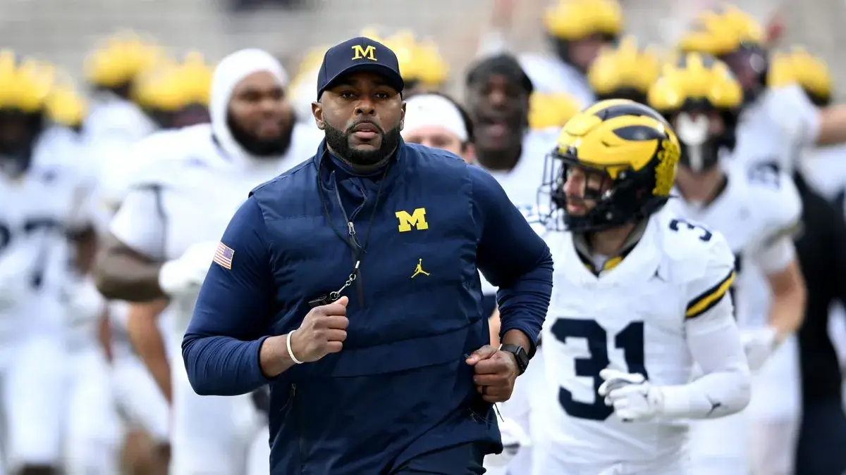 Michigan football coach Sherrone Moore in a navy vest and cap leads his players as they run onto the field before a game.