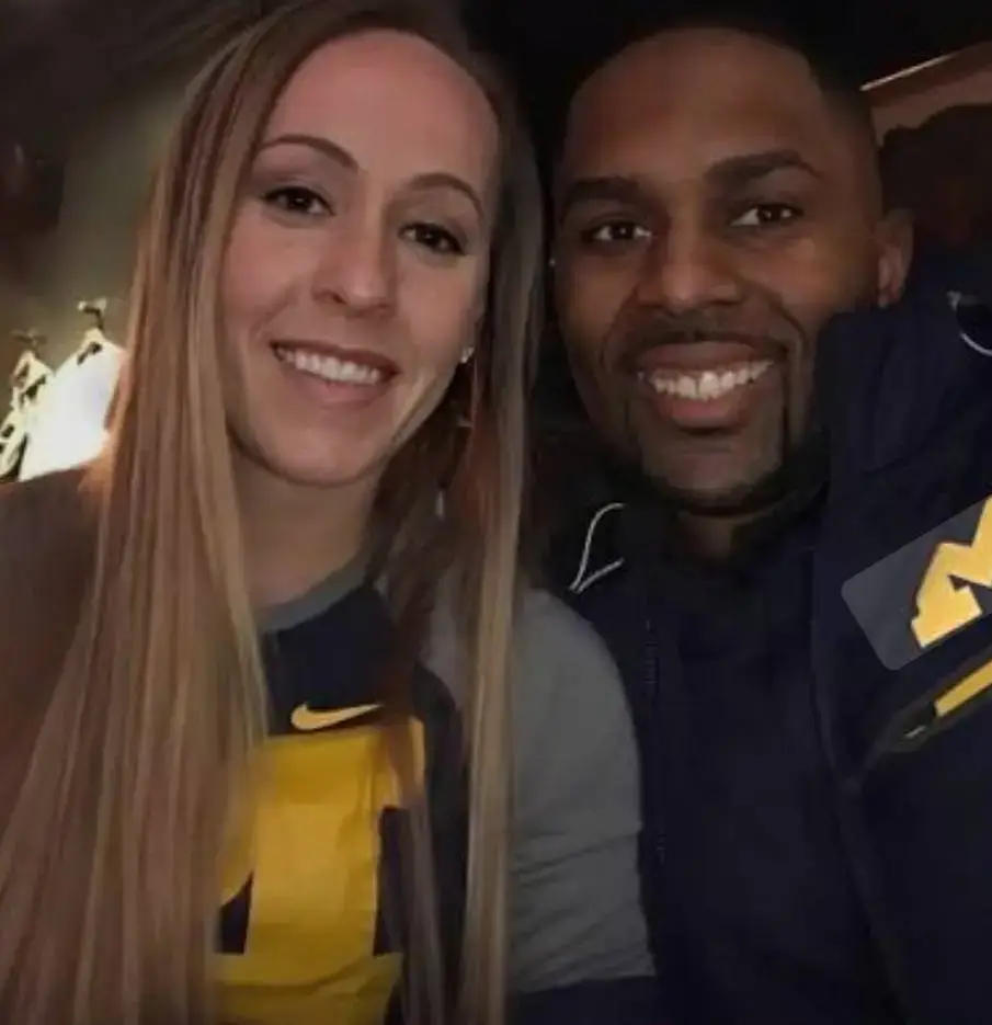 Michigan football coach Sherrone Moore and his wife Kelli Moore smile together in a close-up selfie while wearing casual athletic apparel.