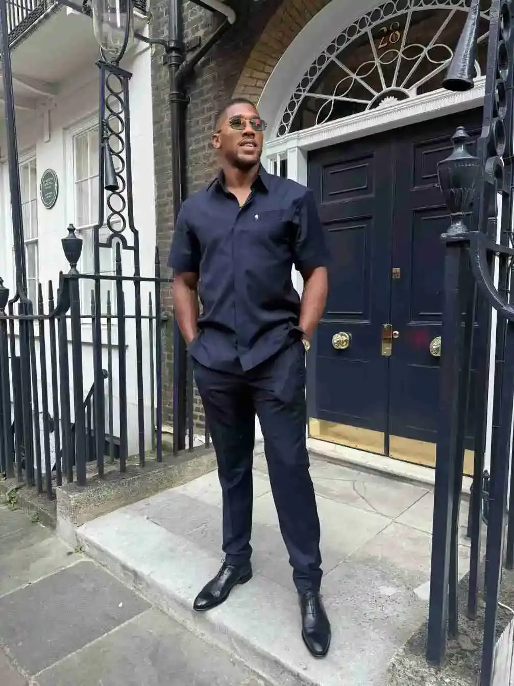 Professional boxer Anthony Joshua stands in front of a dark blue door wearing a navy short-sleeved button-down shirt, matching trousers, and sunglasses with his hands in his pockets.