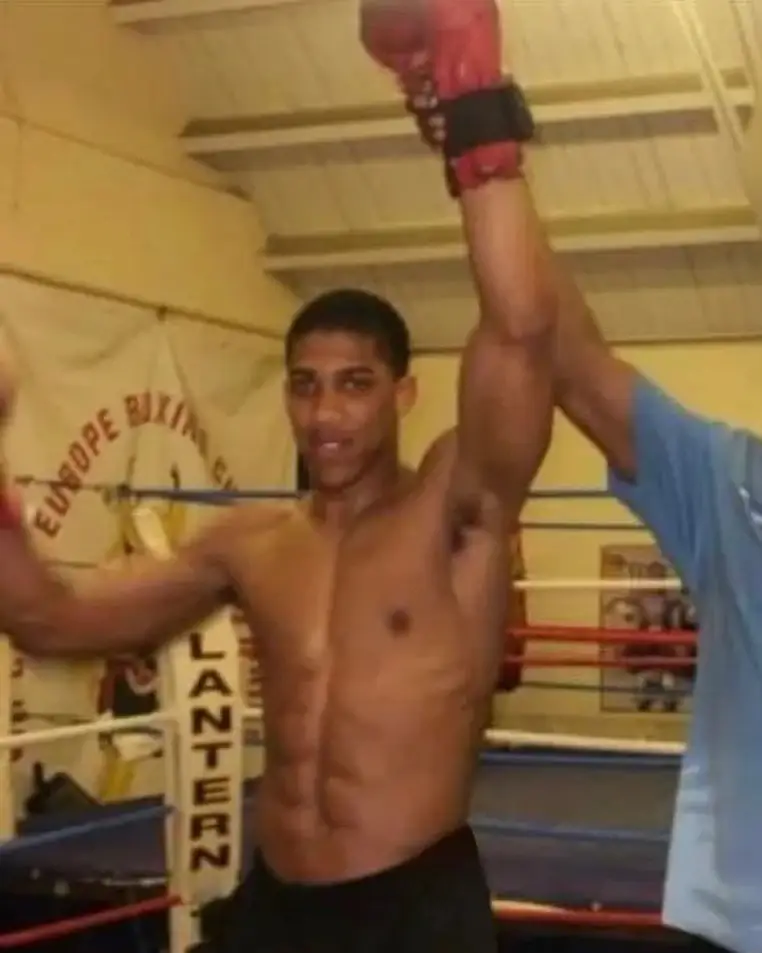 A young Anthony Joshua stands in a boxing ring wearing a red amateur vest and headgear while the referee raises his hand in victory.