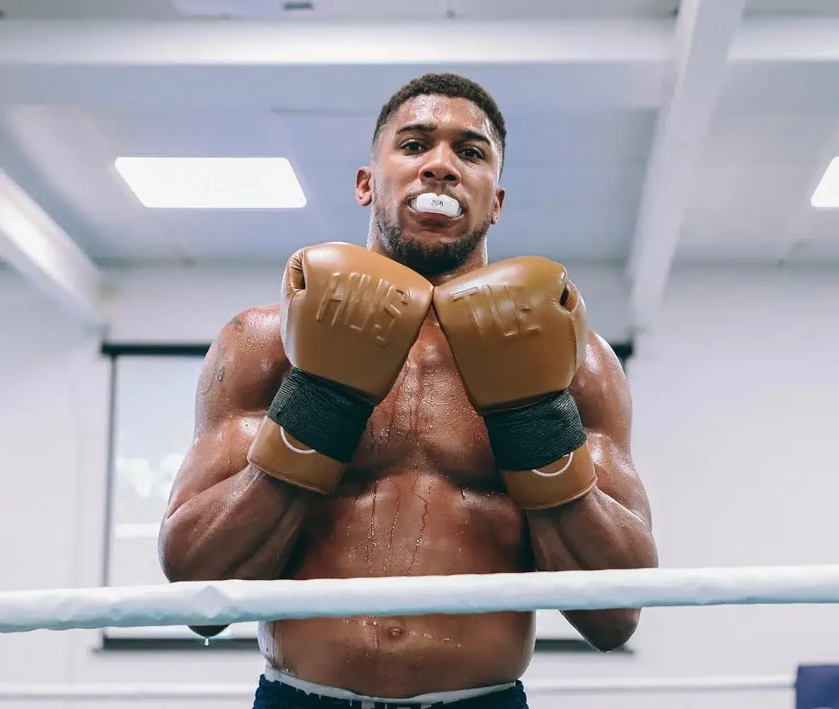 A shirtless and sweaty Anthony Joshua stands in a boxing ring wearing a mouthguard and holding up his brown boxing gloves.