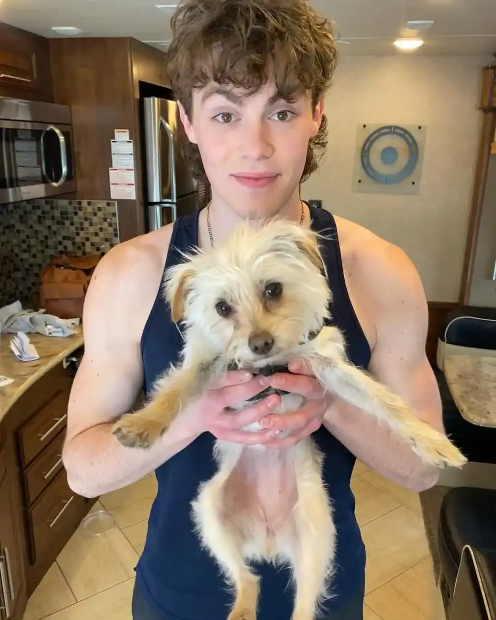 Actor Jack Champion smiles while holding up a small, scruffy cream-colored dog inside a trailer.