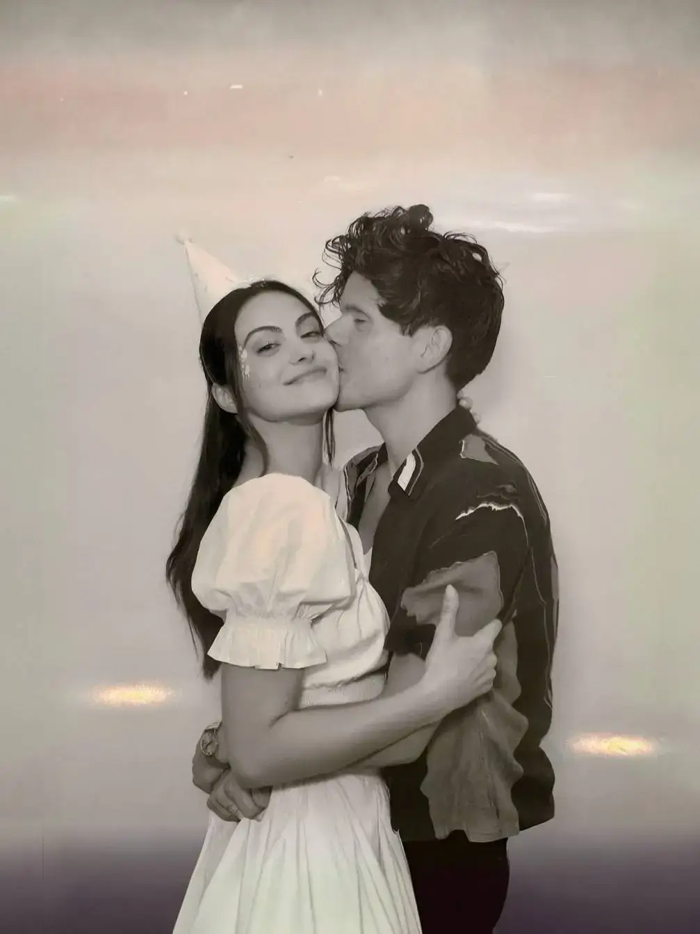 Rudy Mancuso kisses his fiancée Camila Mendes on the cheek as she laughs and wears a party hat in a playful black and white photograph