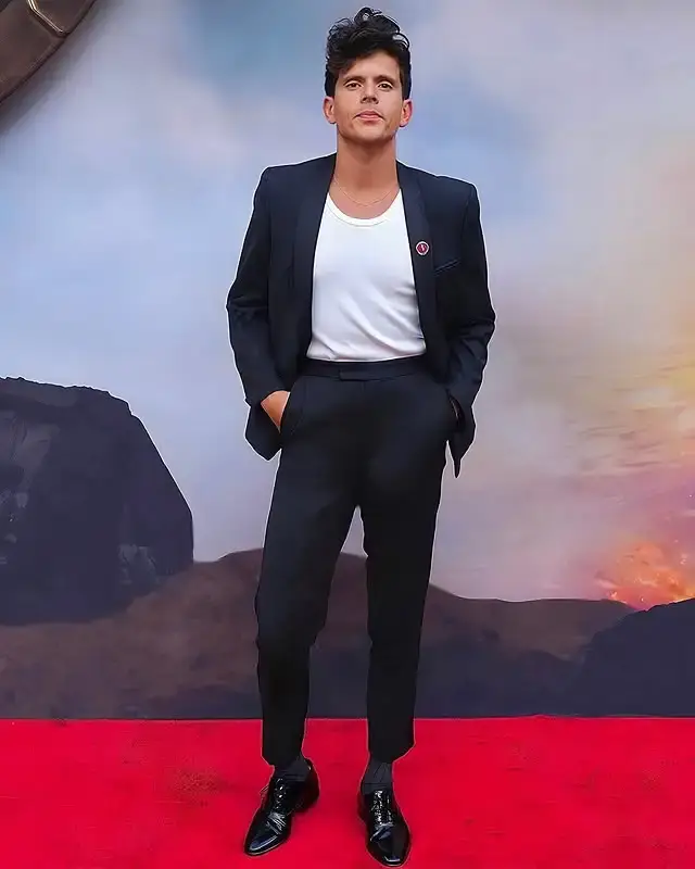 Actor Rudy Mancuso stands on a red carpet with his hands in his pockets, wearing a dark suit over a white t-shirt in front of a volcanic-themed promotional backdrop.