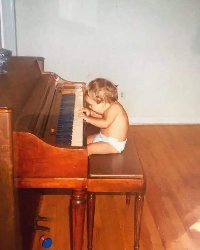 A toddler Rudy Mancuso sits on a piano bench and reaches for the keys of a large wooden upright piano.