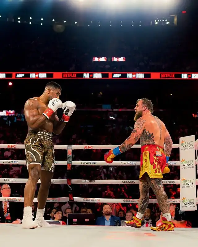 Anthony Joshua, in camouflage trunks, and Jake Paul, in red and yellow flame trunks, face each other in the center of a boxing ring during their match at the Kaseya Center in Miami.