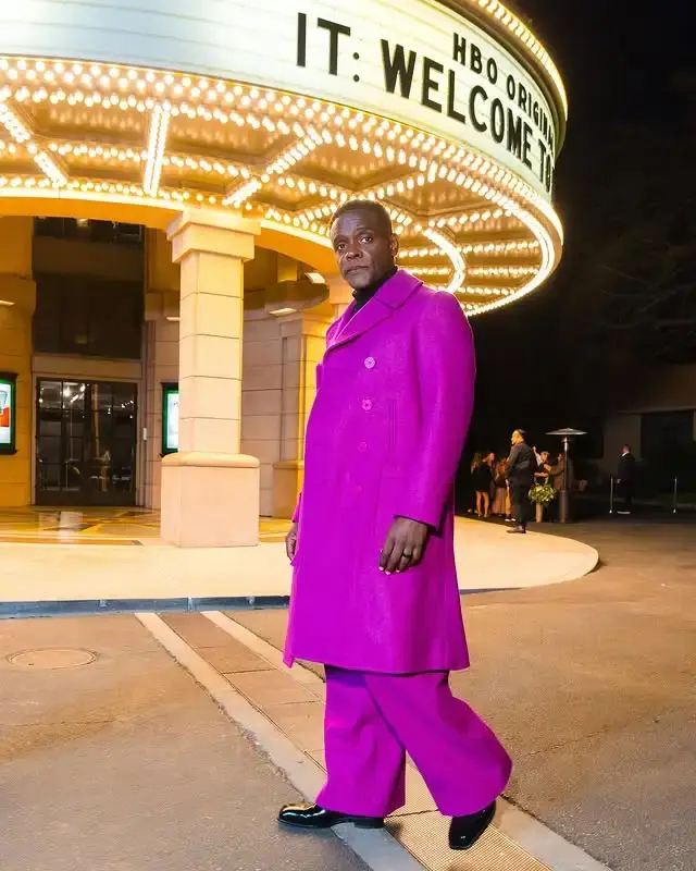 Actor Chris Chalk poses in a vibrant magenta double-breasted coat and matching wide-leg trousers on a sidewalk at night in front of a lit theater marquee for the series "IT: Welcome to Derry".