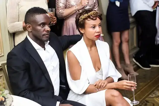 Actor Chris Chalk in a black suit sits beside his wife Kimberley Dalton Mitchell, who wears a white dress and gold headband, at a formal gathering.