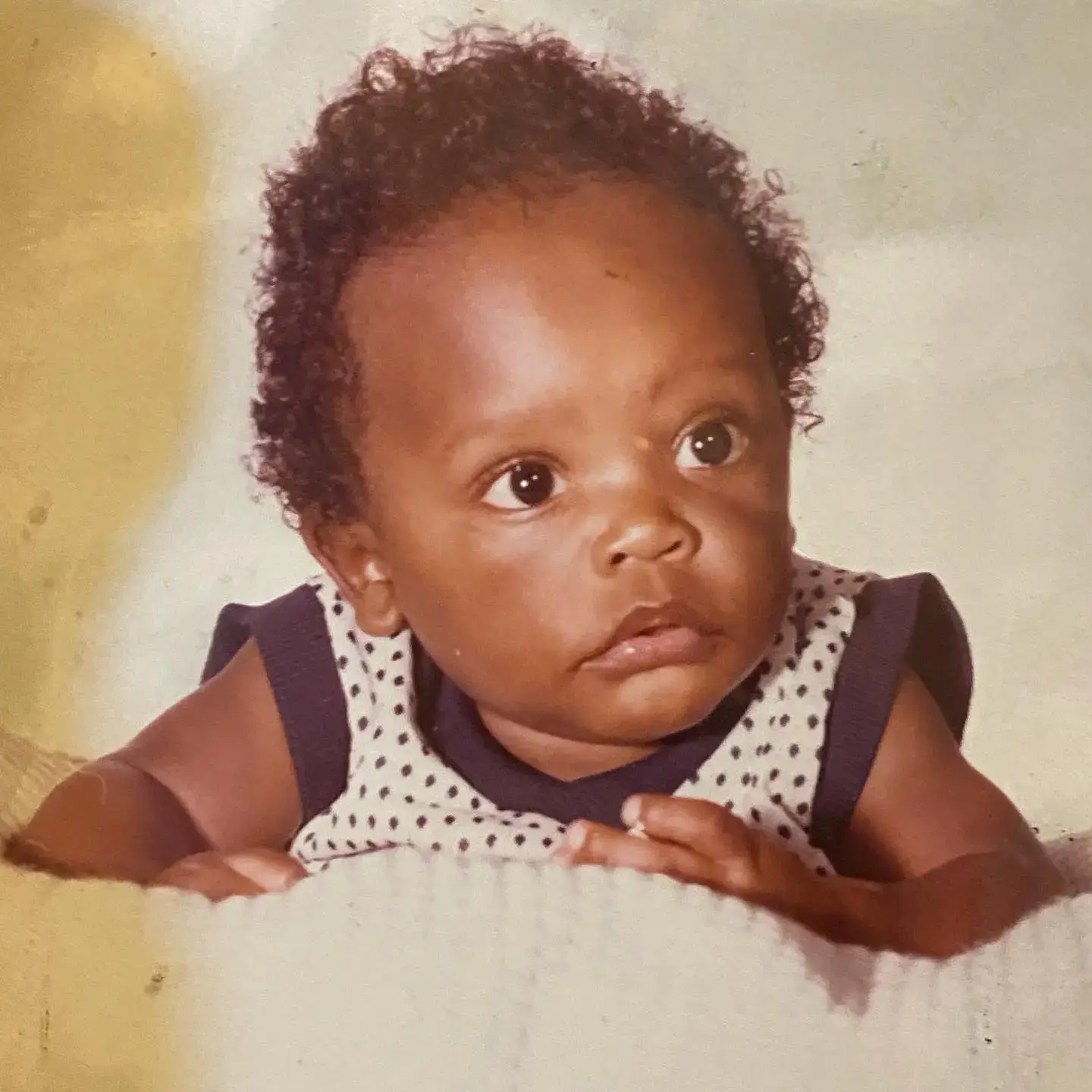 A baby photo of early Chris Chalk with dark curly hair wearing a polka-dot sleeveless shirt while lying on a blanket.