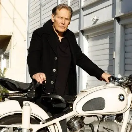 Actor James Remar stands wearing a black pea coat while posing next to a vintage white BMW motorcycle.