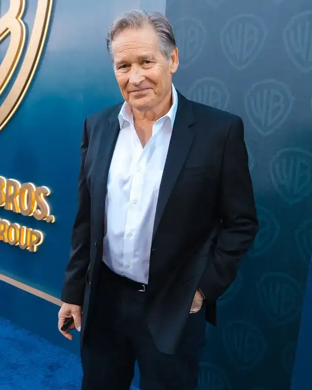 Actor James Remar smiling in a black suit and white dress shirt while standing in front of a blue background with Warner Bros. logos.