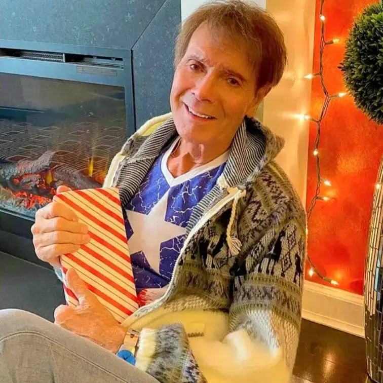 Sir Cliff Richard in this latest picture smiles warmly while sitting by a fireplace and holding a striped gift, dressed in a cozy cardigan and star-print shirt.