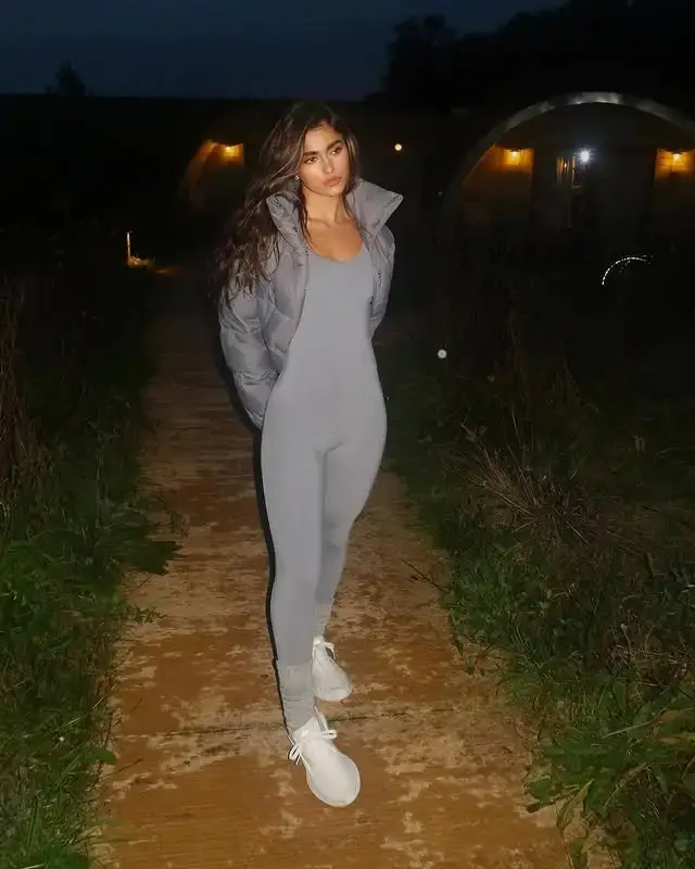 Rayna Vallandingham poses on a wooden path at dusk wearing a sleek grey bodysuit, a matching puffer jacket, and white sneakers.