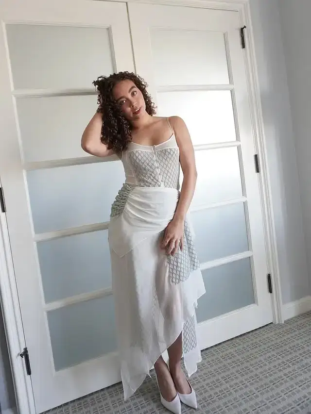 American actress Bailey Bass, known for her role in Avatar: The Way of Water, poses indoors wearing a white sheer dress with grey textured panels.