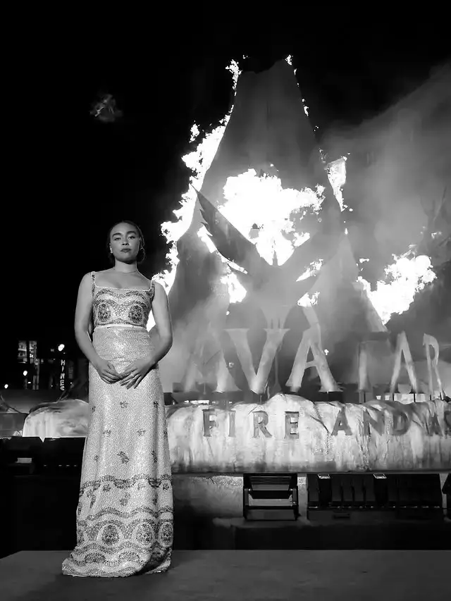 Actress Bailey Bass stands in a shimmering patterned gown in front of a large, fiery Avatar: Fire and Ash promotional display.
