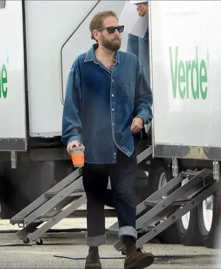 Actor Jonah Hill stands outdoors wearing a blue denim button-down shirt over a white T-shirt, dark sunglasses, and a full beard while holding a beverage cup.