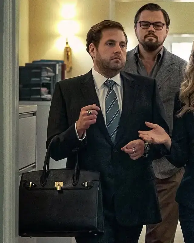 Jonah Hill, dressed in a dark suit and carrying a black Hermès Birkin bag, stands alongside Leonardo DiCaprio in a scene from the film "Don