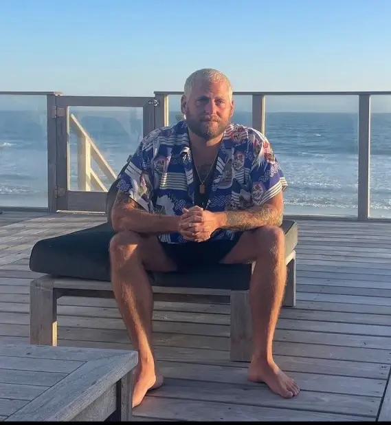 Jonah Hill, with bleached blonde hair and a beard, sits on a wooden lounge chair on a deck overlooking the ocean while wearing a patterned blue button-down shirt.