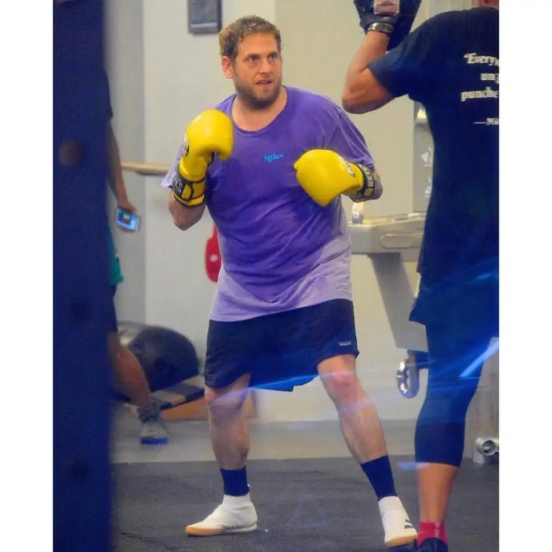 Jonah Hill stands in a boxing ring wearing a purple T-shirt and yellow boxing gloves while maintaining a focused training stance in a gym.