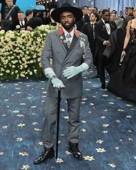 Caleb McLaughlin stands on the 2025 Met Gala red carpet wearing a grey double-breasted Dior suit, turquoise gloves, and a wide-brimmed black hat while holding a silver-handled cane.