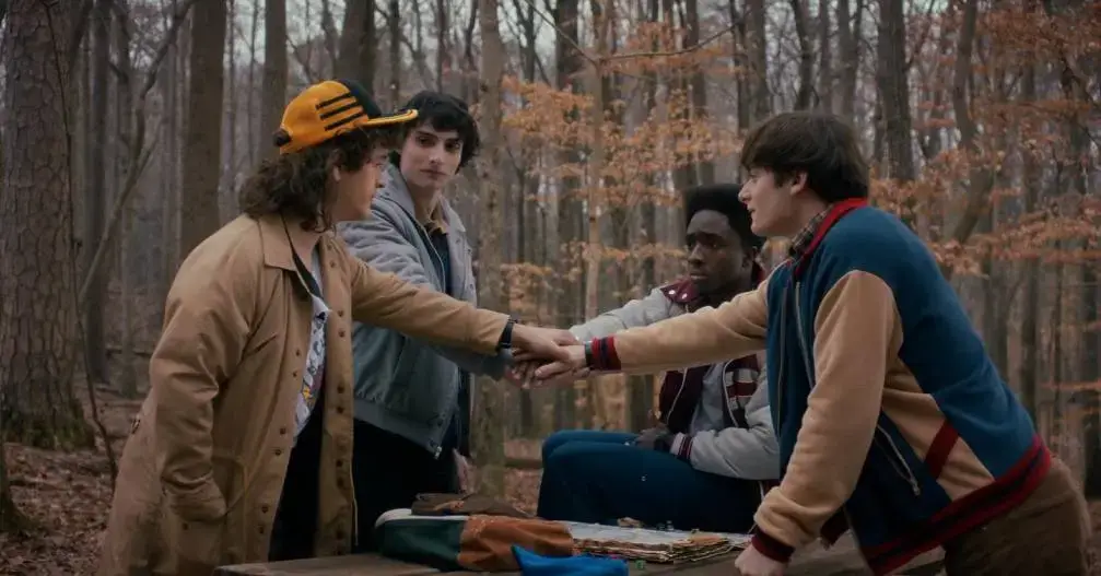 Caleb McLaughlin, Finn Wolfhard, Gaten Matarazzo, and Noah Schnapp sit in a wooded clearing, leaning in to join hands in a central huddle.