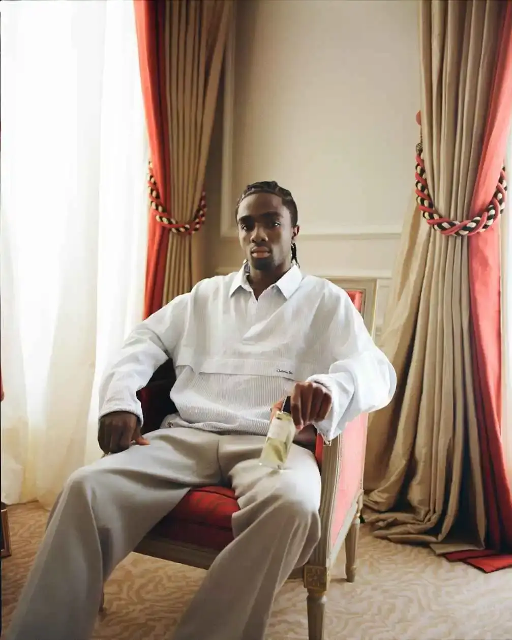 Caleb McLaughlin sits in an ornate red and gold armchair between draped curtains while wearing a white textured long-sleeved shirt and holding a bottle of perfume.