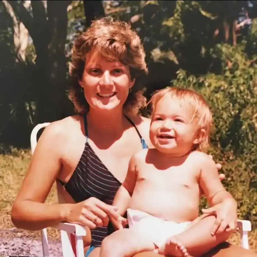 A young Stacy Moore smiles while holding her daughter, Mandy Moore, as a baby on her lap in an outdoor setting.