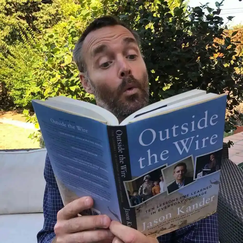 Actor Will Forte makes a surprised facial expression while reading the book "Outside the Wire" by Jason Kander outdoors.