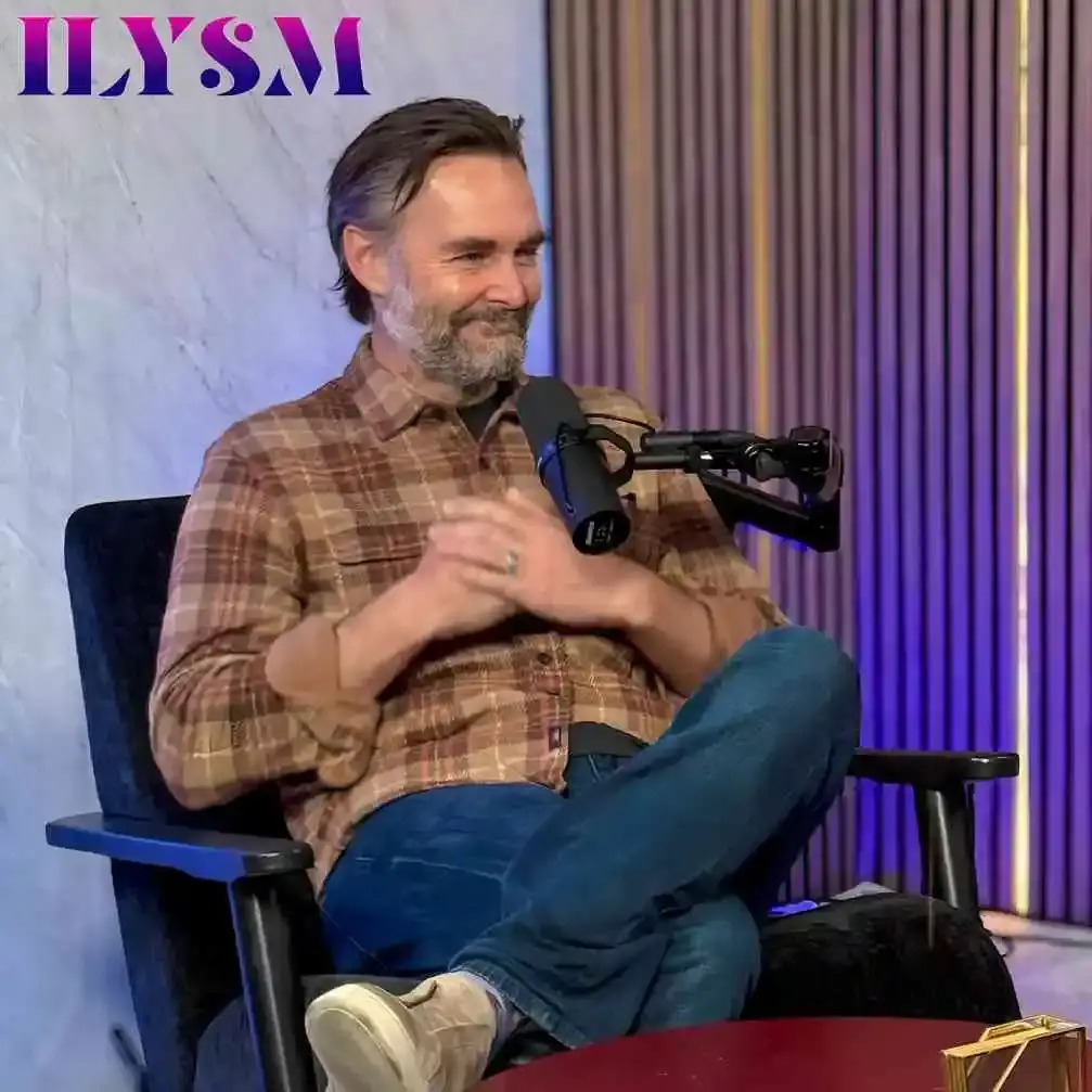 Will Forte sits in a black chair and smiles while wearing a plaid shirt during an interview on the ILYSM podcast.