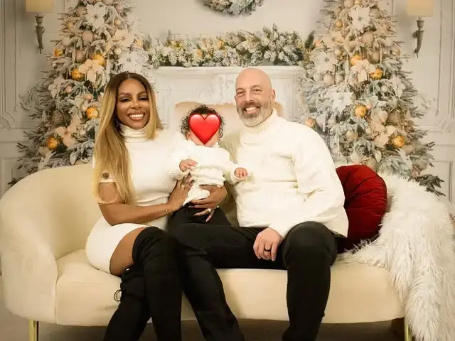 Candiace Dillard Bassett and her husband Chris Bassett pose on a white sofa with their new born baby Jett Maxwell Lee Bassett in front of a festive holiday backdrop featuring decorated Christmas trees.