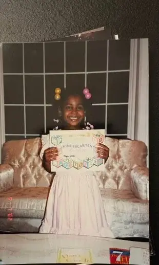 A young Candiace Dillard Bassett smiles brightly while proudly holding up her kindergarten certificate in a vintage childhood photograph.
