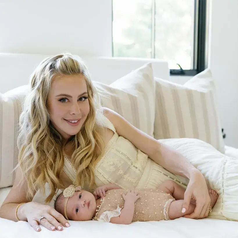 Tara Lipinski smiles and holds her daughter, Georgie Winter, who is dressed in a pink outfit and a matching headband while they pose together outdoors.