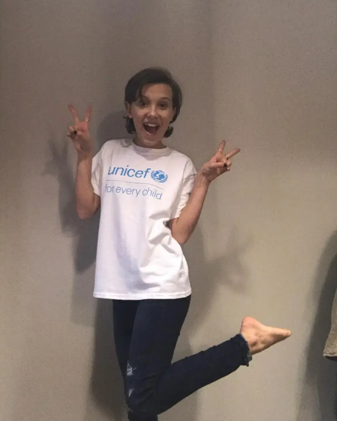 Young Millie Bobby Brown smiles and makes peace signs while wearing a white "unicef for every child" t-shirt.