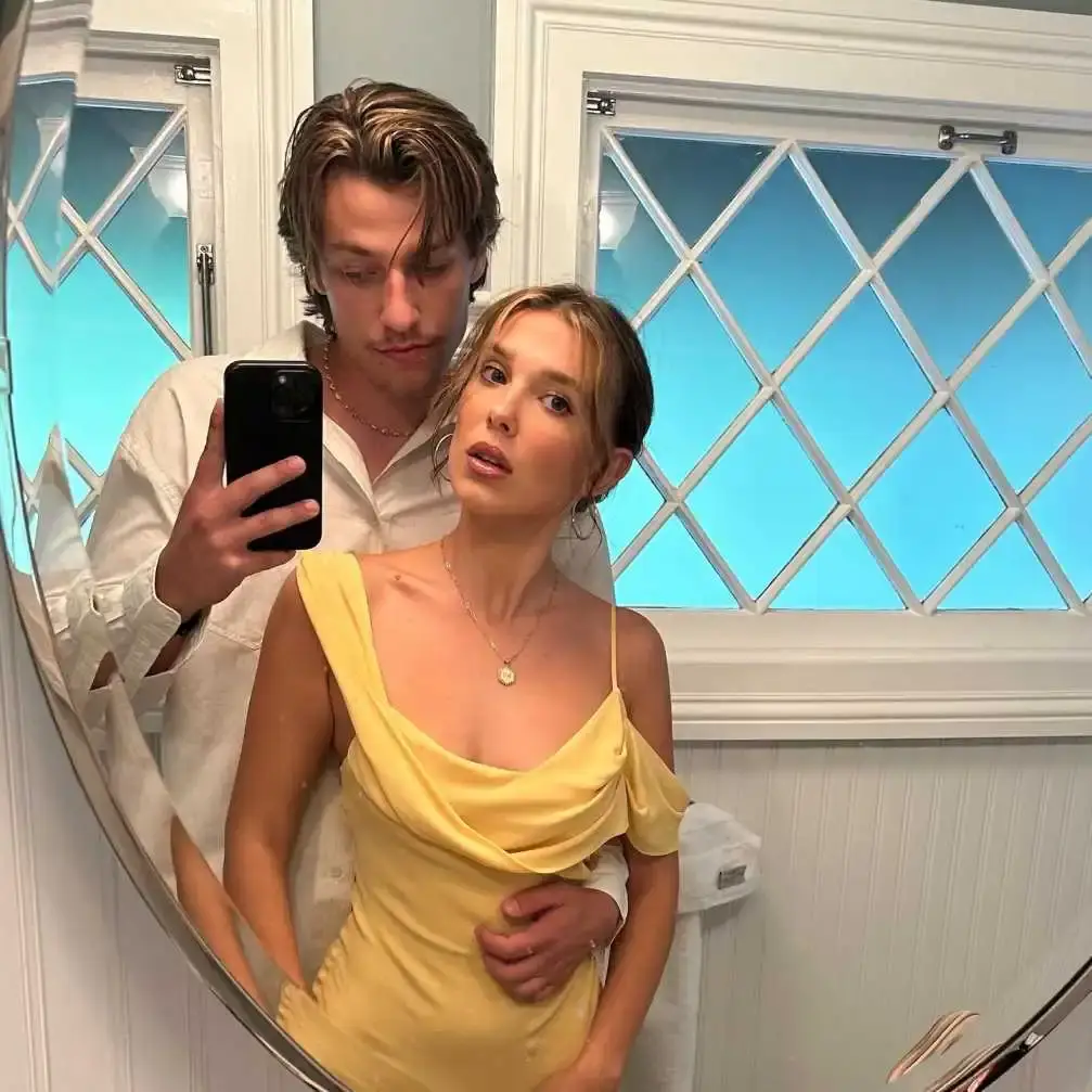 Millie Bobby Brown, wearing a yellow spaghetti-strap dress, poses for a mirror selfie with her husband, Jake Bongiovi, who stands behind her in a white button-down shirt.