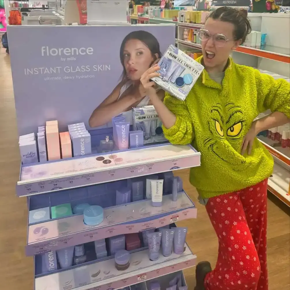 Millie Bobby Brown, wearing a lime green Grinch hoodie and snowflake-patterned pajama pants, smiles excitedly while standing next to a purple and white Florence by Mills retail display.