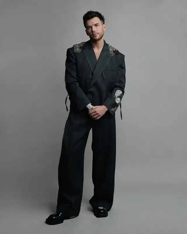 Luke Newton stands against a neutral grey background while wearing a black oversized suit jacket with floral shoulder embellishments and matching wide-leg trousers.