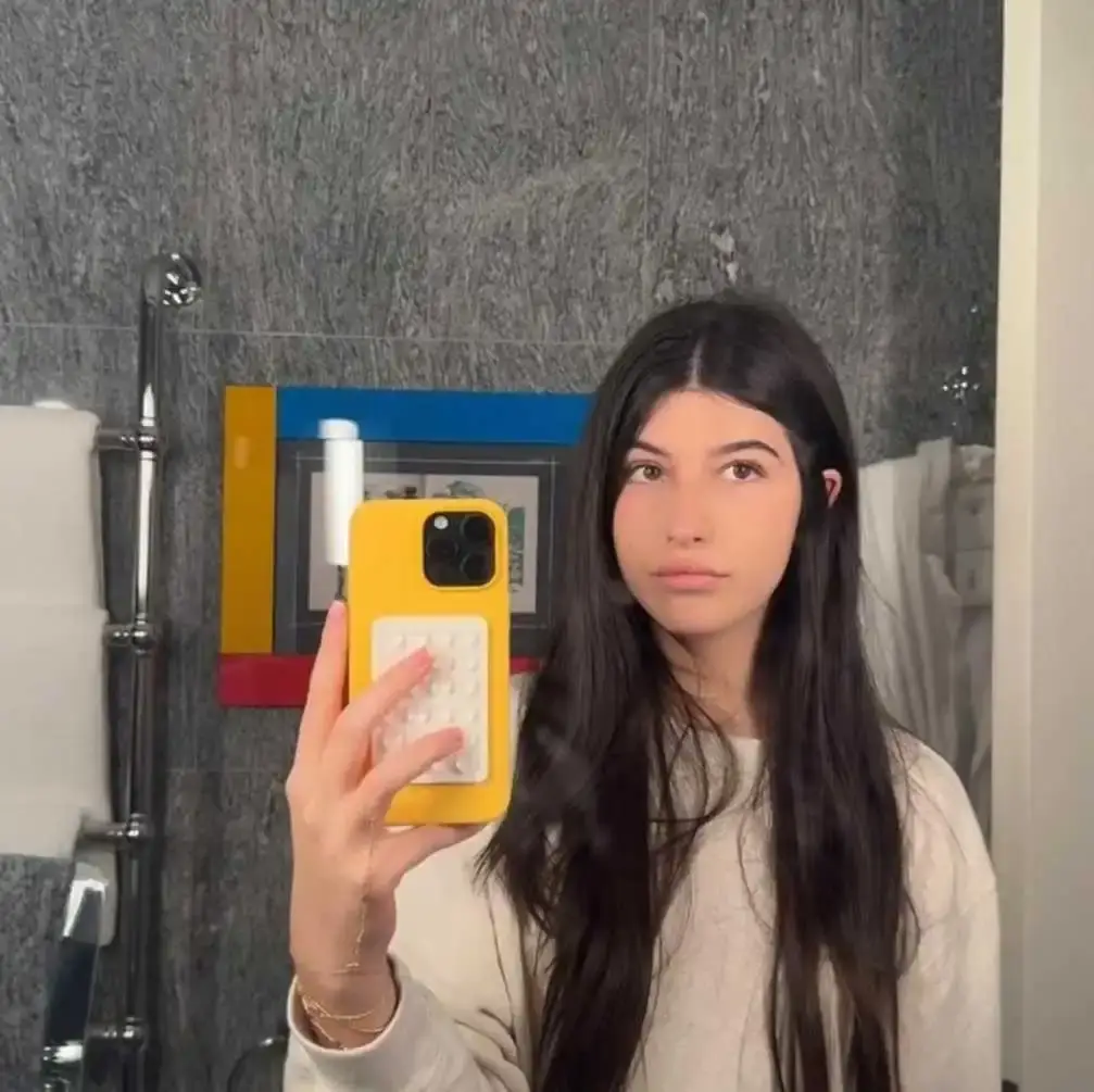 Sunny Sandler takes a mirror selfie in a bathroom while holding a yellow phone with a white pop-it accessory.