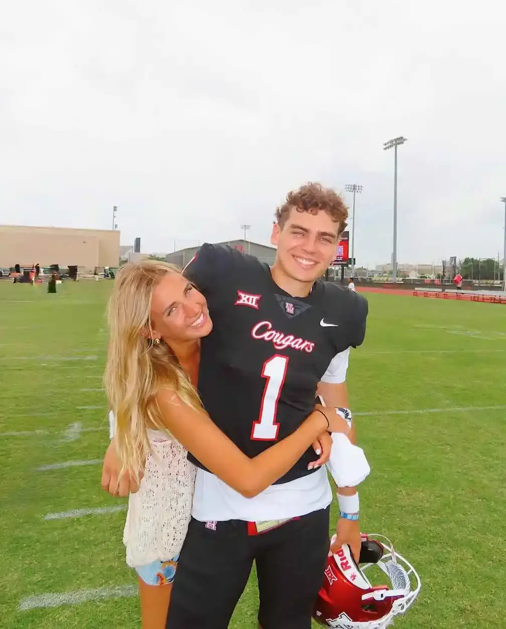 Conner Weigman, wearing a black Houston Cougars football jersey, smiles while being embraced by Hope Griffin on a grass field.