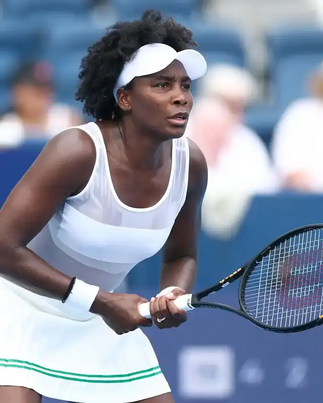 Professional tennis player Venus Williams stands on a court in a white athletic outfit and visor, gripping her racket with both hands in a focused ready position.