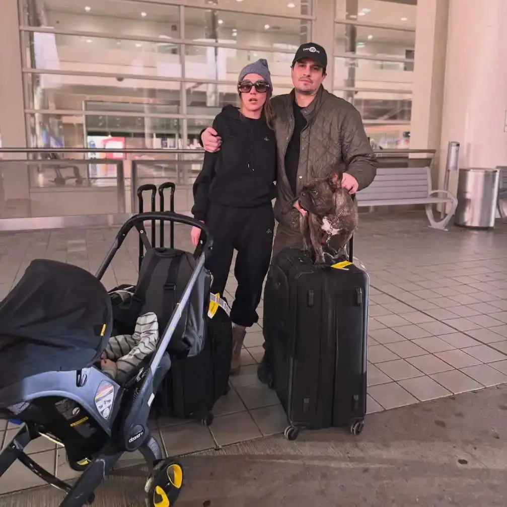 Kat Timpf and Cameron Friscia stand in an airport terminal with their infant son in a stroller and their French Bulldog perched on a suitcase.
