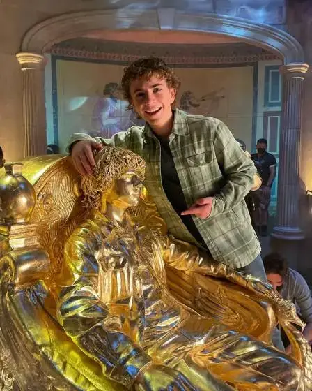 Actor Walker Scobell smiles while leaning on a life-sized golden statue of himself seated in an ornate throne.