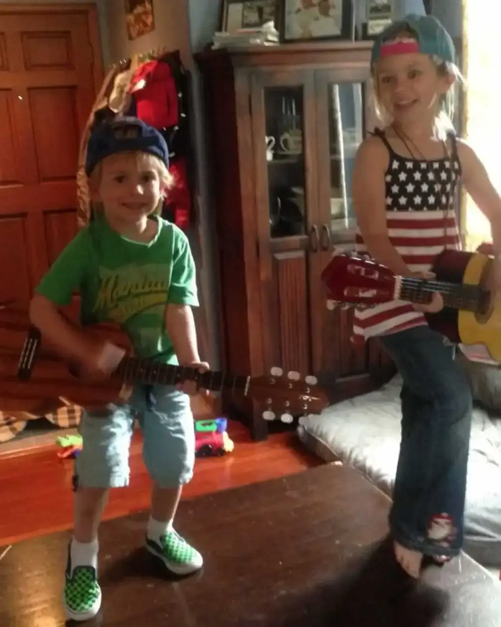 Young siblings Walker Scobell and Leena Scobell stand on a table playing acoustic guitars and wearing backward baseball caps in a childhood home photo.